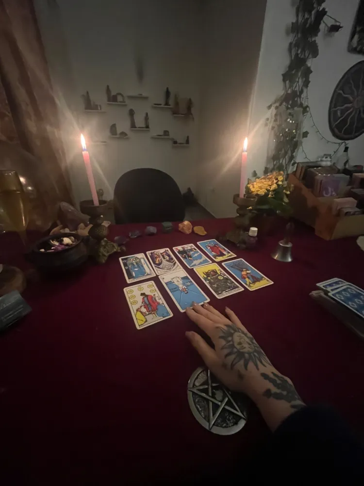 Mão tatuada de Fernanda Verbena sobre uma tiragem de oito cartas de tarot em toalha de veludo vinho, entre velas acesas e cristais